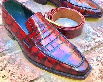 Pode incluir: Um par de mocassins de couro bordô com padrão de crocodilo, combinados com um cinto a condizer com fivela dourada. Os sapatos têm bico quadrado e salto baixo, apresentando um acabamento polido.