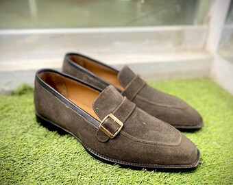 Pode incluir: Um par de mocassins de camurça marrom com uma fivela dourada. Os mocassins estão em uma superfície de grama verde.