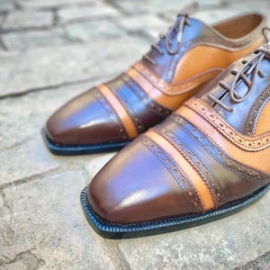 May include: A pair of brown leather shoes with tan and black accents. The shoes have a lace-up closure and a brogue design.