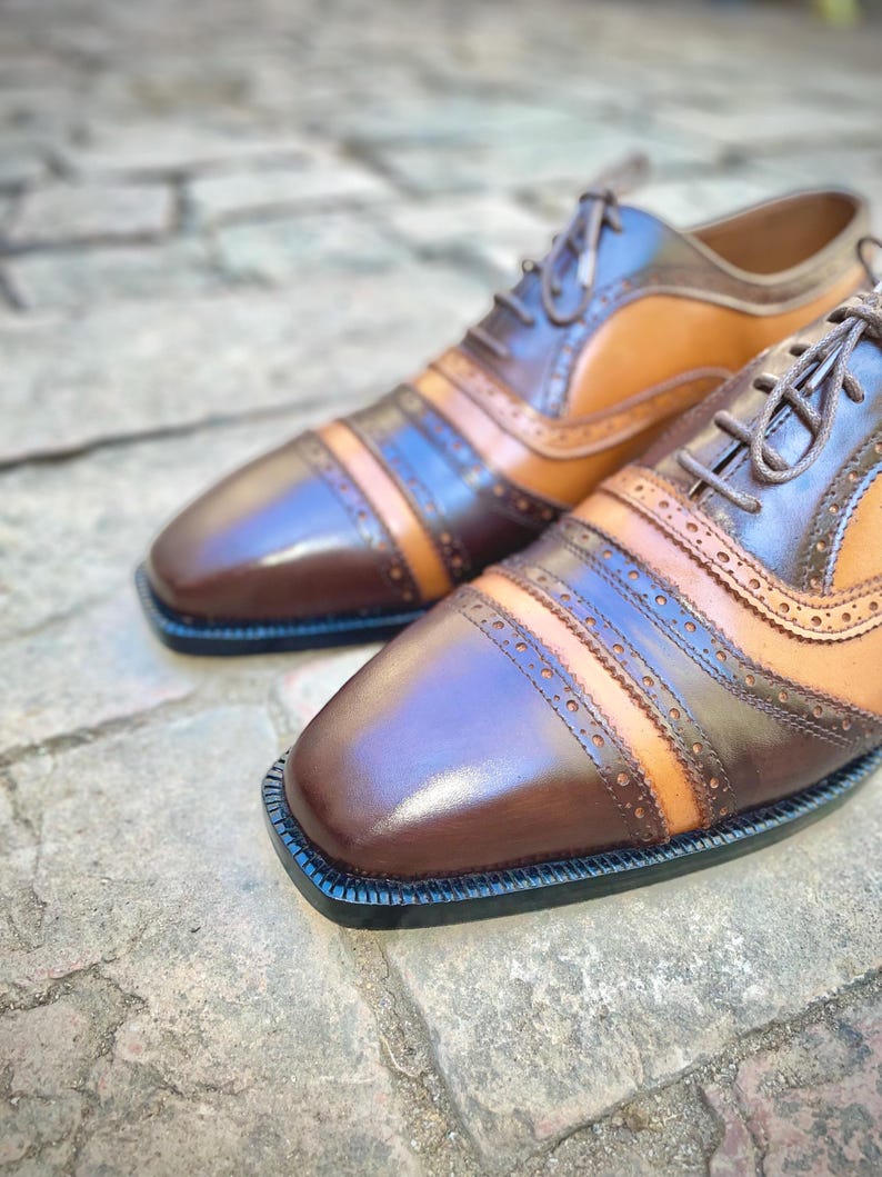 May include: A pair of brown leather shoes with tan and black accents. The shoes have a lace-up closure and a brogue design.