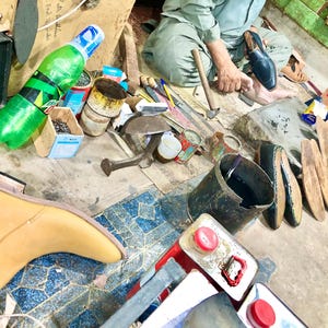 May include: A cobbler's workspace with tools, leather, and a shoe in progress. There are several cans of polish, a bottle of water, and a bucket of black dye. The cobbler is working on a brown leather shoe.