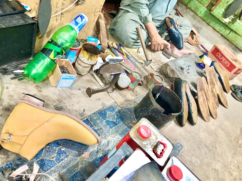 May include: A cobbler's workspace with tools, leather, and a shoe in progress. There are several cans of polish, a bottle of water, and a bucket of black dye. The cobbler is working on a brown leather shoe.
