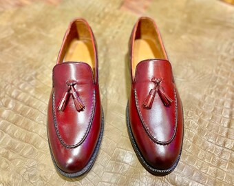 Pode incluir: Um par de mocassins de couro bordô com detalhes de borlas. Os sapatos têm bico arredondado e sola preta. O interior é de cor bege claro. Os mocassins são exibidos em uma superfície texturizada marrom claro.