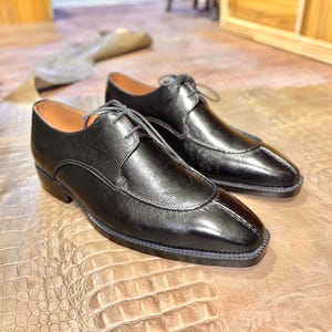 May include: A pair of black leather dress shoes with lace-up closures. The shoes have a pointed toe and a slightly squared-off shape. The shoes are sitting on a brown leather surface.