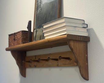 Puede incluir: Estante de madera rústica con acabado natural, con una foto de paisaje enmarcada, una cesta tejida y una pila de libros. Debajo, hay seis clavijas para colgar objetos. El estante está montado en una pared blanca.