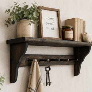 May include: A dark brown wooden wall shelf with a flat top surface and a lower rail featuring five metal hooks. The shelf holds a potted green plant, a framed sign reading "HOME sweet HOME", a glass candle jar, three books, and a small bird figurine.