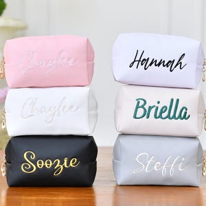 May include: A stack of six personalized cosmetic bags in various colors: pink, white, black, light gray, and off-white. Each bag has a name embroidered in a contrasting color, with gold-tone zipper pulls. The names include "Chaylee", "Hannah", "Briella", "Soozie", and "Steffi".