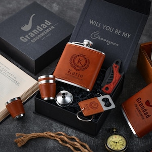 May include: A gift set featuring a brown leather-wrapped flask, shot glasses, and a bottle opener. The flask is engraved with a monogram and the name "Katie". The set includes a pocket knife and is presented in a black box with the text "Will you be my Groomsmen".