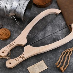May include: Two wooden cheese slicers with stainless steel blades, each engraved with a name. One is labeled "Callum" and the other "Kevin". The slicers are light-colored wood and have a curved handle design. A small wooden tag with "1891" is also visible.