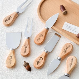 May include: A collection of cheese knives and forks with wooden handles and stainless steel blades. Each handle is engraved with a name. The set includes a variety of blade shapes and fork styles, perfect for serving cheese.