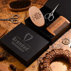 May include: A grooming kit with a black box featuring the text "Grandad Groomsman". The kit includes a wooden comb, brush, scissors, and a wooden oval brush with the text "Grandad Groomsman Est. 2020". The items are arranged on a map.