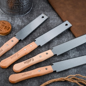 Könnte beinhalten: Vier kleine Handsägen mit Holzgriffen, jede mit einem anderen Namen graviert: Mikey, Jahnee, Caitlin und Morgan. Die Sägeblätter sind silberfarben und die Griffe sind helles Holz. Die Sägen sind auf einer dunklen Oberfläche angeordnet.