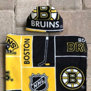 May include: A black, yellow, and white fleece hat and blanket set. The hat and blanket feature the Boston Bruins logo and the words "BRUINS" and "BOSTON" in yellow lettering. The blanket also has the NHL logo.