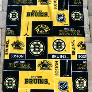 May include: A black, yellow, and white Boston Bruins fleece blanket. The blanket features the team's logo, the words "BOSTON BRUINS", and hockey stick graphics. The NHL logo is also visible.