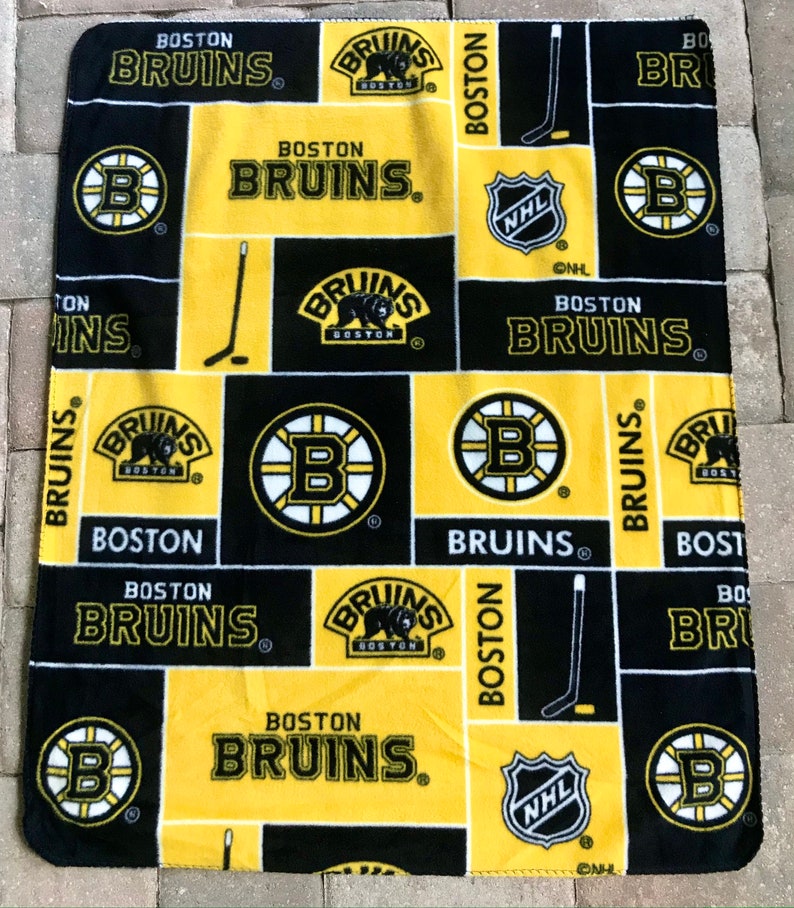 May include: A black, yellow, and white Boston Bruins fleece blanket. The blanket features the team's logo, the words "BOSTON BRUINS", and hockey stick graphics. The NHL logo is also visible.