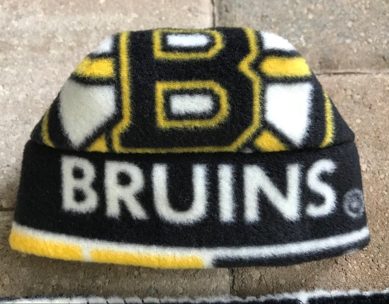 May include: A black, yellow, and white fleece beanie hat. The hat features the word "BRUINS" in white block letters across the front. The top of the hat has a repeating pattern of the letter "B" in black and yellow.