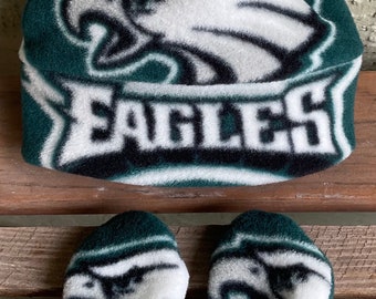 Puede incluir: Un conjunto de gorro y mitones de forro polar verde y blanco. El gorro presenta la palabra "EAGLES" y un logotipo de cabeza de águila estilizada. Los mitones y el gorro son verdes con detalles en blanco y negro.