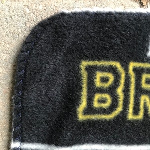 May include: A black and gold blanket with the word "BR" in yellow block letters. The blanket has a white stripe and a dark blue stitched edge. The blanket is made of a soft, fuzzy material.