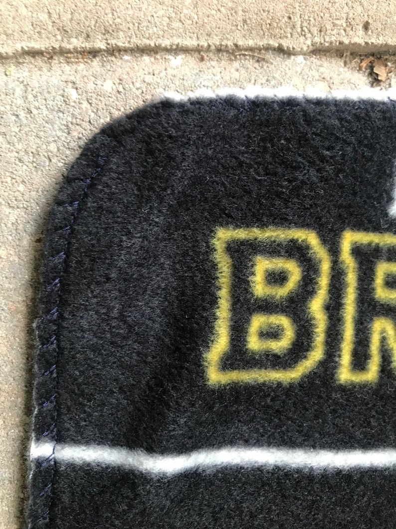 May include: A black and gold blanket with the word "BR" in yellow block letters. The blanket has a white stripe and a dark blue stitched edge. The blanket is made of a soft, fuzzy material.