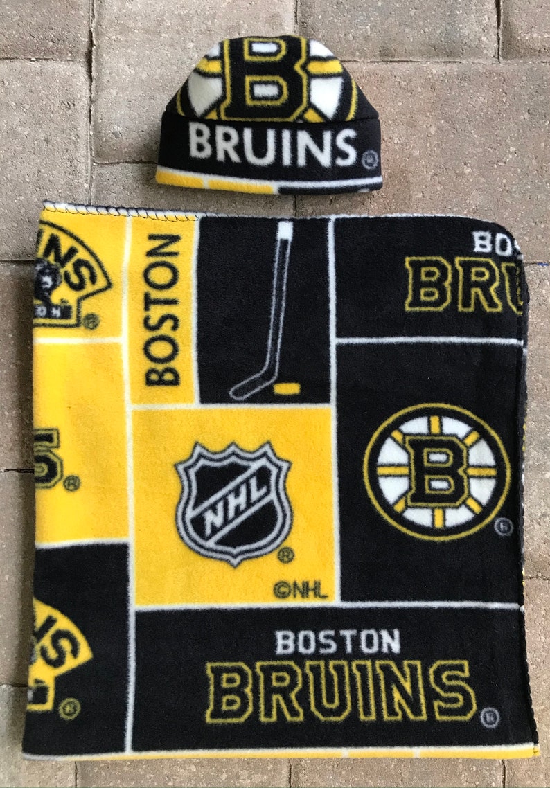 May include: A black, yellow, and white fleece hat and blanket set. The hat and blanket feature the Boston Bruins logo and the words "BRUINS" and "BOSTON" in yellow lettering. The blanket also has the NHL logo.