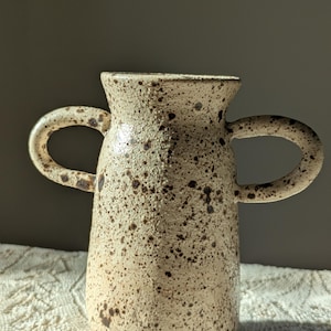May include: A speckled ceramic vase with two loop handles. The vase has a cream-coloured base with brown speckles. The handles are rounded and attached to the upper portion of the vase. The vase is sitting on a white, textured surface.