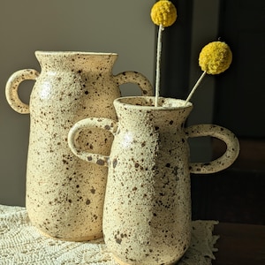 May include: Two speckled ceramic vases with loop handles. The vases are beige with brown speckles. One vase is taller than the other. The smaller vase holds two yellow pom-pom flowers. The vases are on a cream-coloured lace doily.