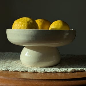 May include: A cream-coloured ceramic pedestal bowl holding three bright yellow lemons. The bowl has a wide, shallow top and a rounded base. The lemons are the focal point, contrasting with the neutral tones of the bowl and background.