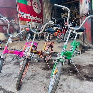 Puede incluir: Tres bicicletas vintage con manillares y marcos cromados. Las bicicletas son de varios colores, incluyendo rosa, verde y marrón. Las bicicletas están aparcadas en un garaje.