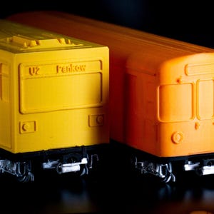 May include: Two toy train carriages, one yellow and one orange, are set side-by-side against a black backdrop. The yellow carriage has the text "U2 Pankow" on the front. The trains have detailed undercarriages.