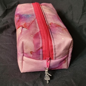 May include: A rectangular, pink cosmetic bag with a watercolor design in shades of pink, purple, and red. The bag has a bright red zipper and a silver ballerina charm. The bag is made of fabric.
