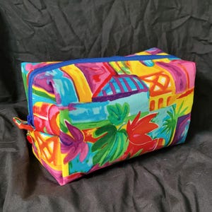 May include: A colorful rectangular zippered pouch with a bright abstract pattern of houses, flowers, and leaves. The pouch is made of fabric and has a blue zipper.
