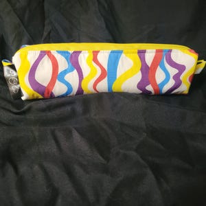 May include: A rectangular pencil case with a white background and a pattern of wavy lines in yellow, red, purple, and blue. The zipper is yellow, and there is a small tag on the side. The case is on a black surface.