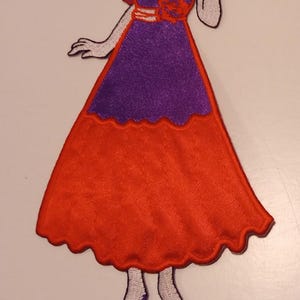 May include: An embroidered fabric patch depicting a figure wearing a purple bodice and a long orange skirt with a scalloped hem, a red hat, and a gold necklace. A wooden ruler placed at the base shows the figure is approximately 4 inches tall.
