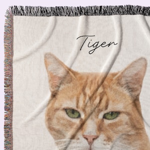 Puede incluir: Una manta de color crema con un primer plano de la cara de un gato atigrado naranja con ojos verdes. La palabra "Tiger" est&aacute; escrita en cursiva. La manta tiene un borde con flecos.