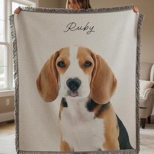 Puede incluir: Una manta tejida beige con un retrato de un perro beagle. El perro tiene pelaje marr&oacute;n y blanco y ojos azules. El nombre "Ruby" est&aacute; escrito en cursiva en la parte superior. La manta tiene un borde con flecos.