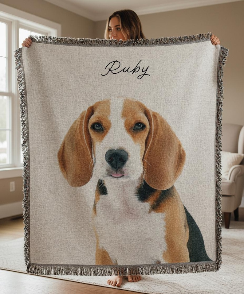 Puede incluir: Una manta tejida beige con un retrato de un perro beagle. El perro tiene pelaje marr&oacute;n y blanco y ojos azules. El nombre "Ruby" est&aacute; escrito en cursiva en la parte superior. La manta tiene un borde con flecos.