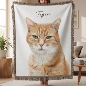 Puede incluir: Una manta crema con un retrato grande y detallado de un gato atigrado naranja con ojos verdes. La palabra "Tiger" est&aacute; escrita en cursiva. La manta tiene un borde con flecos.
