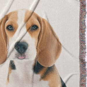 Puede incluir: Una manta tejida con un retrato detallado de un perro Beagle. El perro tiene marcas marrones y blancas, una nariz negra y orejas ca&iacute;das. La manta tiene un borde con flecos multicolores.