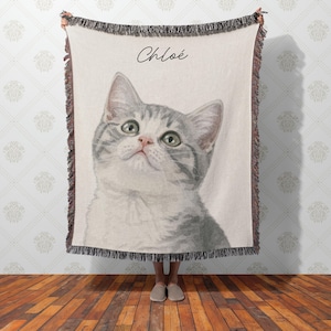 Puede incluir: Una manta crema con un retrato de estilo acuarela de un gato atigrado gris y blanco con ojos verdes. El gato est&aacute; mirando hacia arriba. El nombre "Chlo&eacute;" est&aacute; escrito en cursiva sobre la cabeza del gato. La manta tiene un borde con flecos.