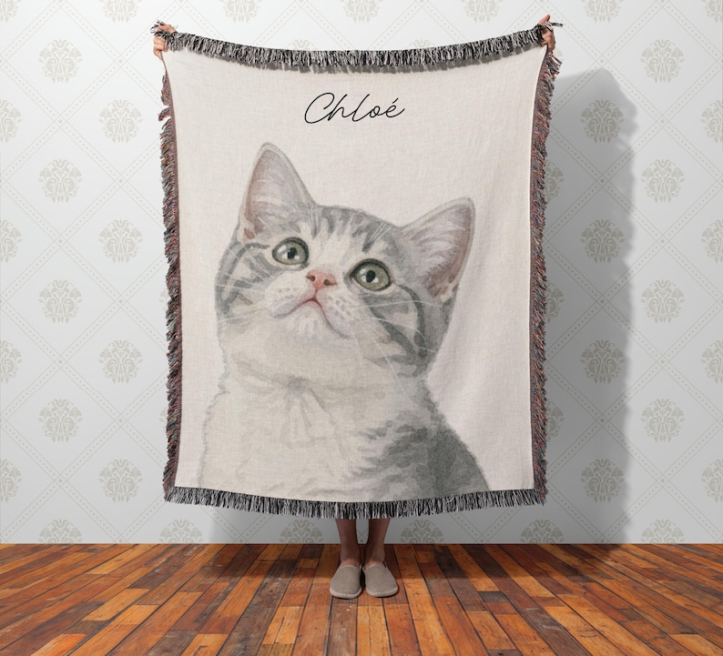 Puede incluir: Una manta crema con un retrato de estilo acuarela de un gato atigrado gris y blanco con ojos verdes. El gato est&aacute; mirando hacia arriba. El nombre "Chlo&eacute;" est&aacute; escrito en cursiva sobre la cabeza del gato. La manta tiene un borde con flecos.