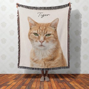 Puede incluir: Una manta tejida con un retrato detallado de un gato atigrado naranja con ojos verdes. La palabra "Tiger" est&aacute; escrita en cursiva sobre la cabeza del gato. La manta tiene un borde con flecos.