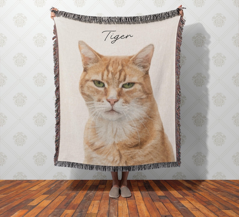 Puede incluir: Una manta tejida con un retrato detallado de un gato atigrado naranja con ojos verdes. La palabra "Tiger" est&aacute; escrita en cursiva sobre la cabeza del gato. La manta tiene un borde con flecos.