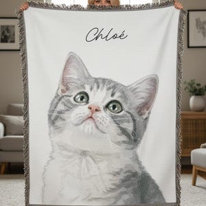 Puede incluir: Una manta blanca con una ilustraci&oacute;n de acuarela de un gato gris y blanco con ojos verdes. El nombre "Chlo&eacute;" est&aacute; escrito en cursiva encima del gato. La manta tiene un borde con flecos.