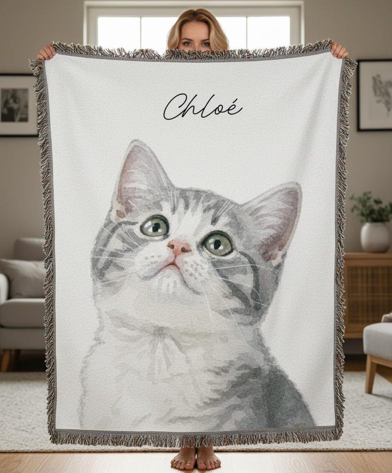 Puede incluir: Una manta blanca con una ilustraci&oacute;n de acuarela de un gato gris y blanco con ojos verdes. El nombre "Chlo&eacute;" est&aacute; escrito en cursiva encima del gato. La manta tiene un borde con flecos.