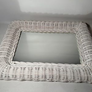 May include: A rectangular white wicker mirror. The frame is made of woven wicker and surrounds a reflective glass surface. The mirror is a decorative item, suitable for various interior design styles.