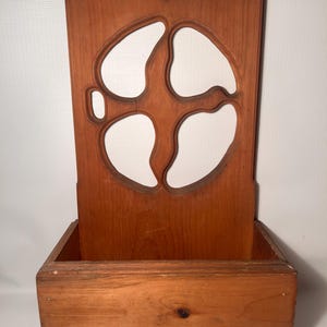 Könnte beinhalten: Wandhalterung für Streichhölzer und Reiber aus Holz. Der Halter besteht aus gebeiztem Holz mit einem dekorativen Ausschnitt. Der Boden hat eine kleine Schachtel für Streichhölzer. Die Oberseite hat ein Loch zum Aufhängen.