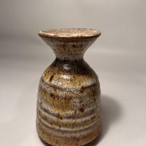 May include: A small, handcrafted ceramic vase with a flared top and a rounded body. The vase has a textured surface with horizontal bands of brown, white, and yellow hues. The design is rustic and earthy.