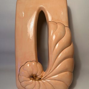 May include: A peach-coloured ceramic wall sculpture with a rectangular frame and a central arched opening. The lower right features a stylised, spiralling seashell design with gold accents. The sculpture has a glossy finish.