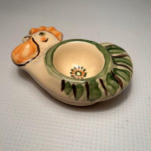Può includere: Una ciotola decorativa in ceramica a forma di gallo. La ciotola è color crema con accenti arancioni, verdi e marroni. Il gallo ha un volto dettagliato e una ciotola circolare sulla schiena, con un piccolo motivo floreale sul fondo.