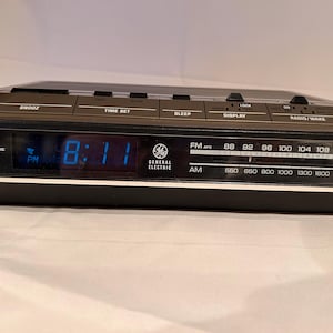 Puede incluir: Un reloj despertador digital negro General Electric con una pantalla digital azul que muestra las 8:11 PM. El reloj tiene botones para repetición, ajuste de hora, dormir, visualización y radio/despertador. El dial de radio muestra las frecuencias FM y AM.